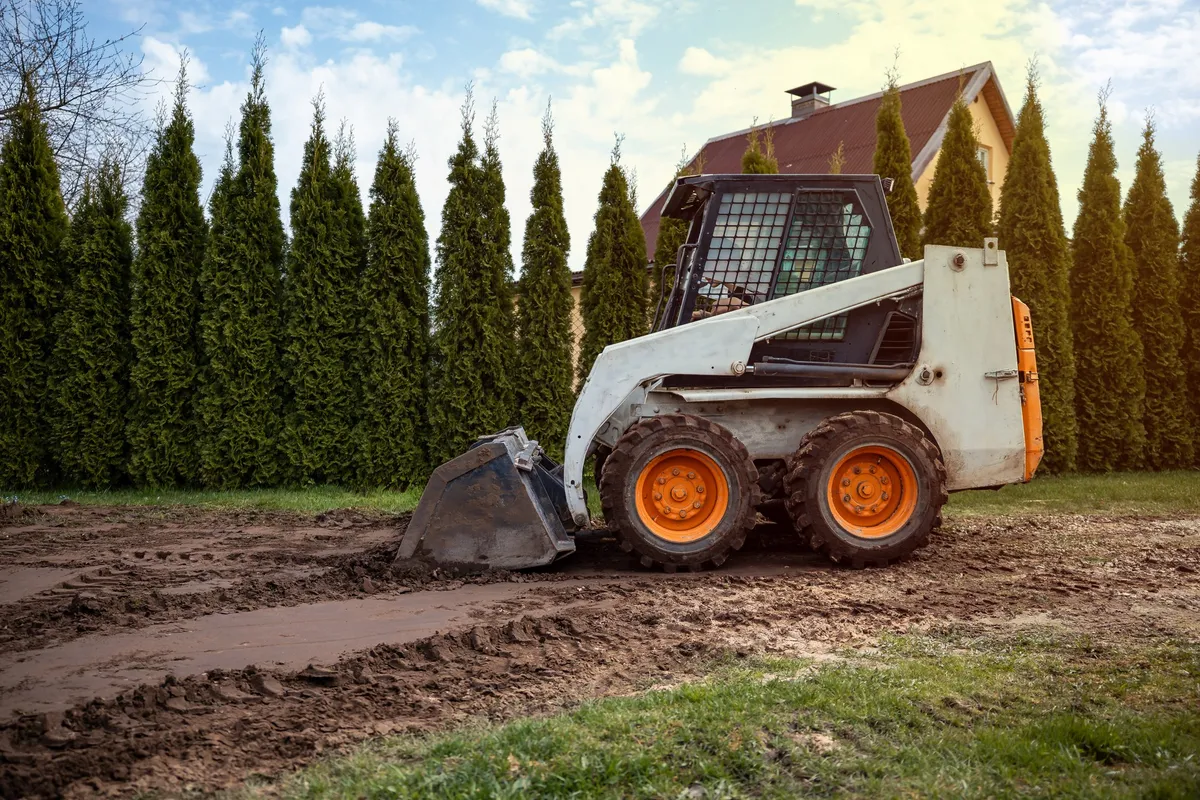 A mini skid steer loader on a dirt path surrounded by evergreen trees and a house, enhancing job efficiency.